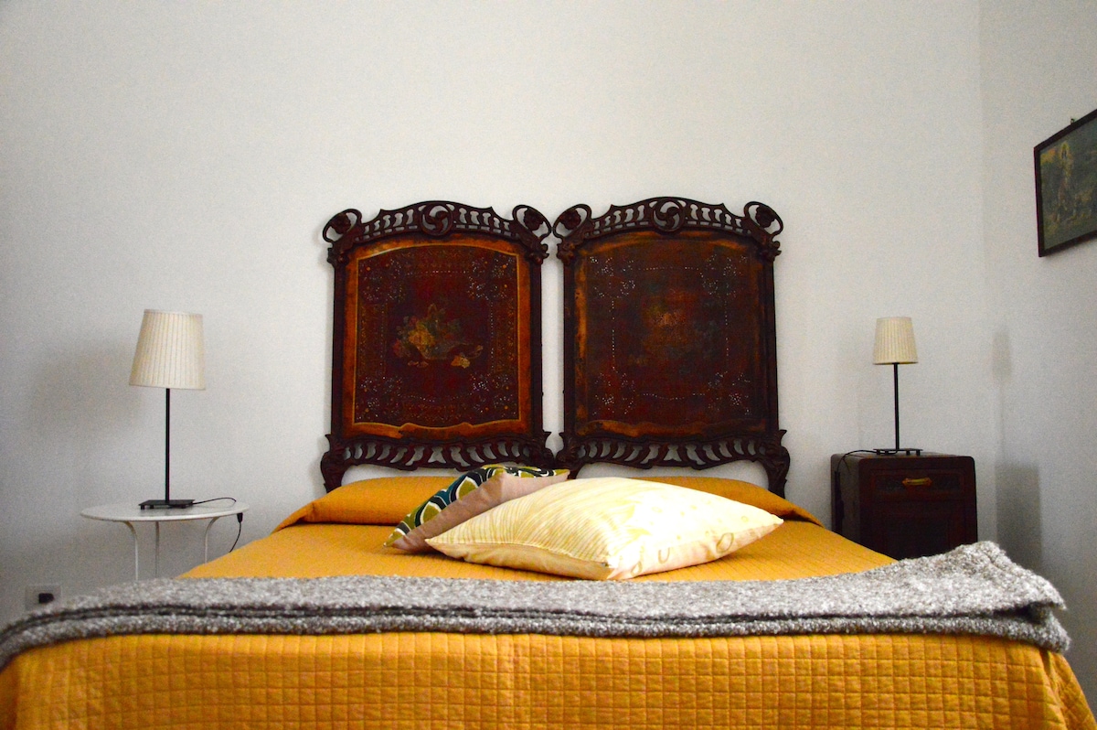 A spacious bedroom features an intricately carved headboard with detailed artwork. Two bedside lamps provide soft light, and light yellow bedding complements the neutral walls. A gray throw blanket is casually draped across the bed, adding a layer of texture to the welcoming space.