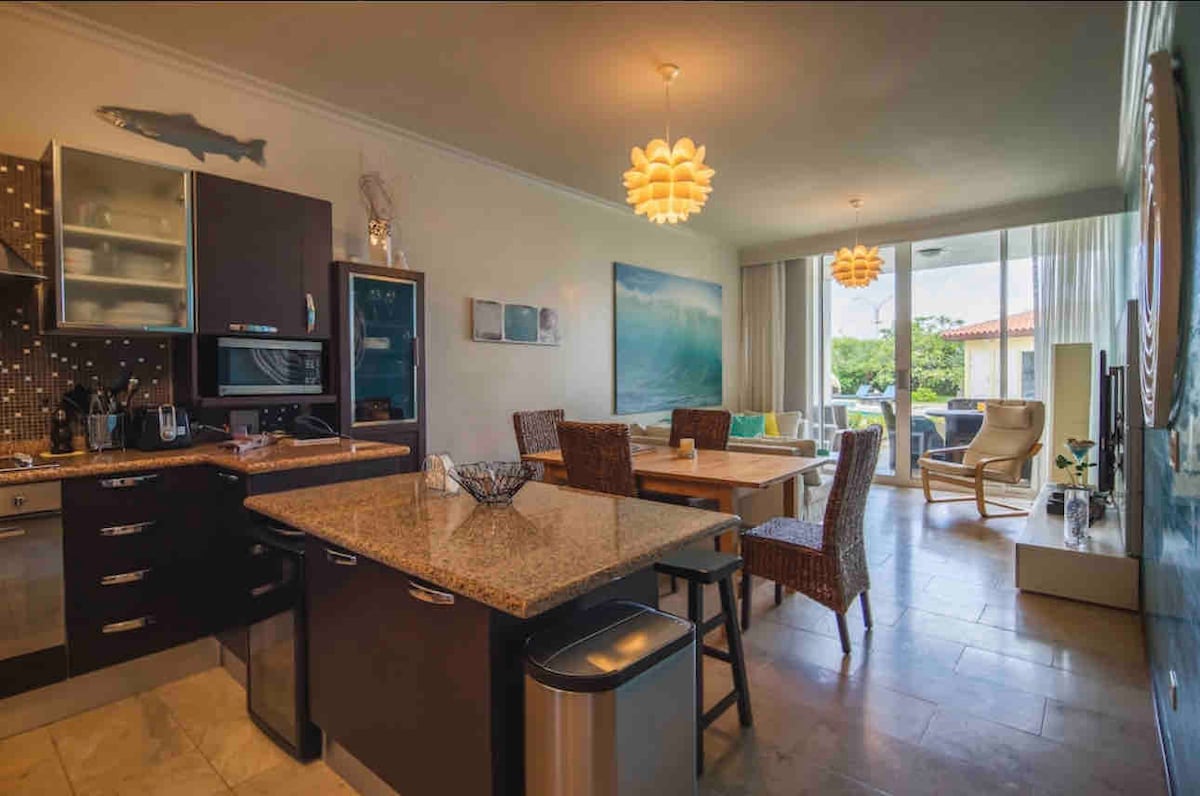 An open kitchen and living room area features contemporary dark cabinetry and a large granite countertop. A dining table with woven chairs is positioned by expansive windows, allowing natural light to fill the space. A serene artwork depicting water adorns the wall.