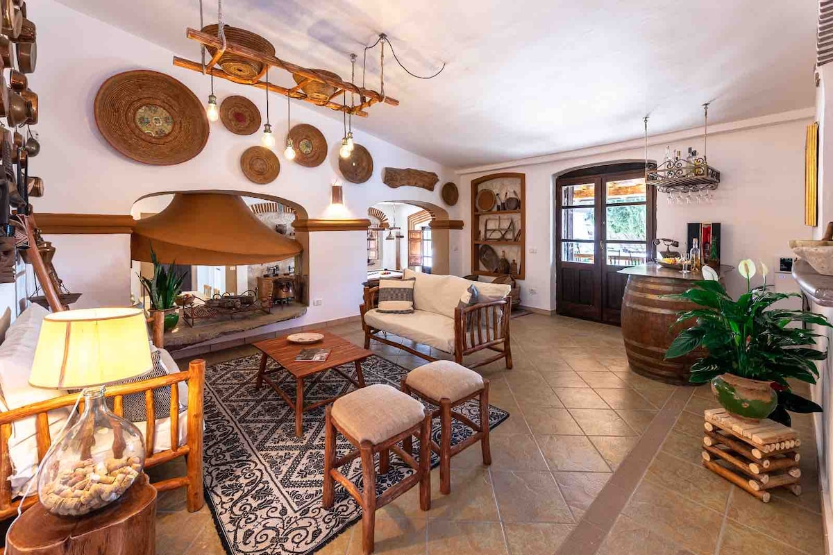 The living area features a traditional fireplace centered under a wooden beam ceiling. Rustic decor, including woven baskets, adorns the walls. A light-colored sofa and two wooden stools are arranged around a low table, creating a cozy atmosphere. Large windows allow natural light to fill the space.