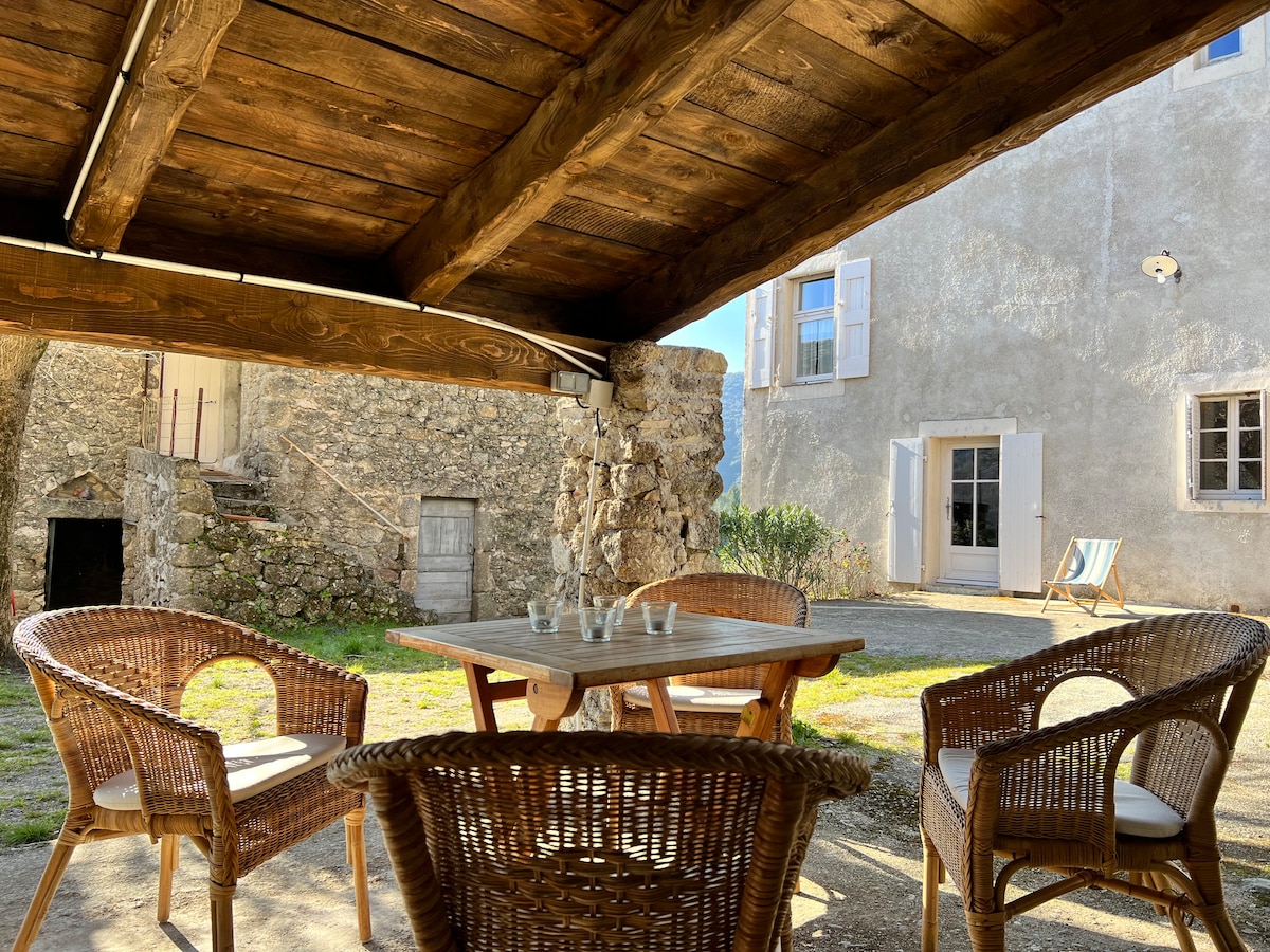 An outdoor seating area is set under a wooden structure, featuring a table surrounded by woven chairs. The backdrop includes a stone wall and the white exterior of the house, with windows allowing natural light to fill the space.