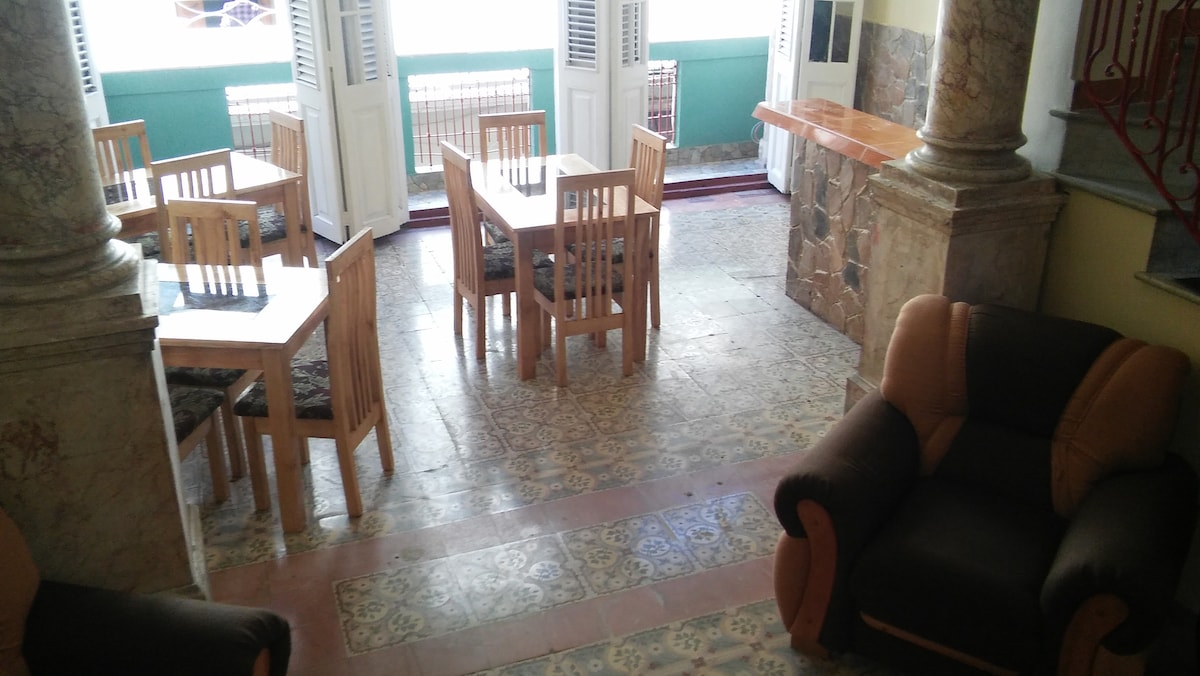 A dining area features several wooden tables surrounded by matching chairs, creating a comfortable space for meals. Pillared architecture frames the open space, which is enhanced by colorful tiled flooring. Large windows allow natural light to fill the room, revealing a decorative bar area.