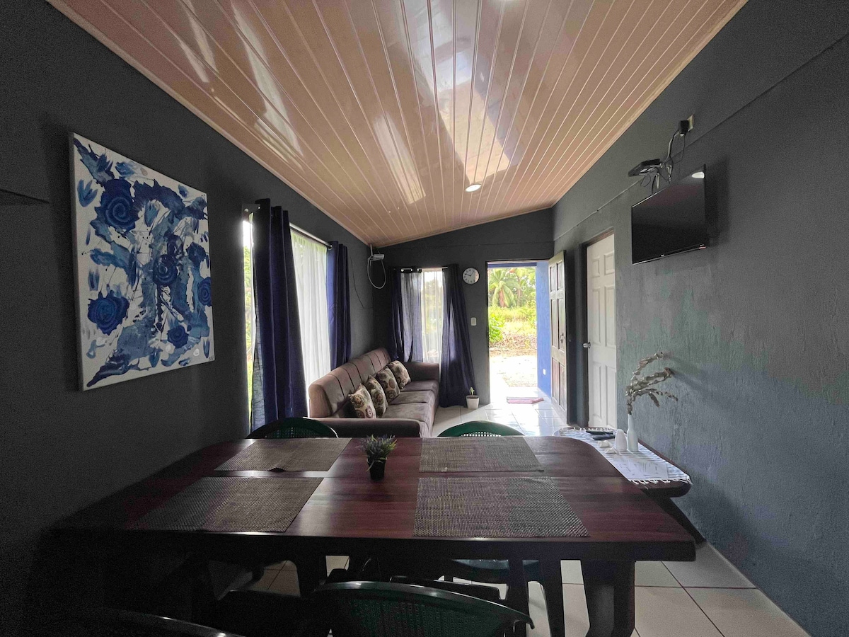 A spacious living area is featured, with large windows letting in natural light. A comfortable sofa is positioned against a wall, complemented by a dining table with green chairs. Wall art adds a touch of color, and a television is mounted for entertainment.