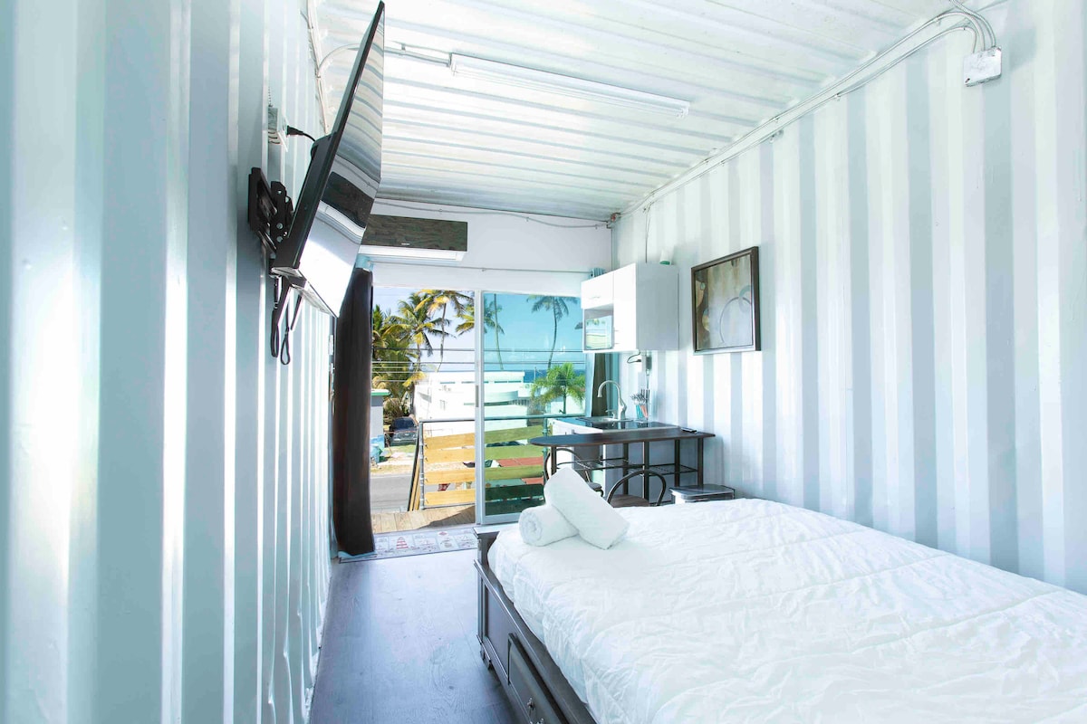 A bright sleeping area is feature with a spacious bed topped with a white comforter. A mounted television is positioned on the wall. A desk and chair are situated nearby, while large sliding glass doors invite natural light and provide access to an outdoor space.