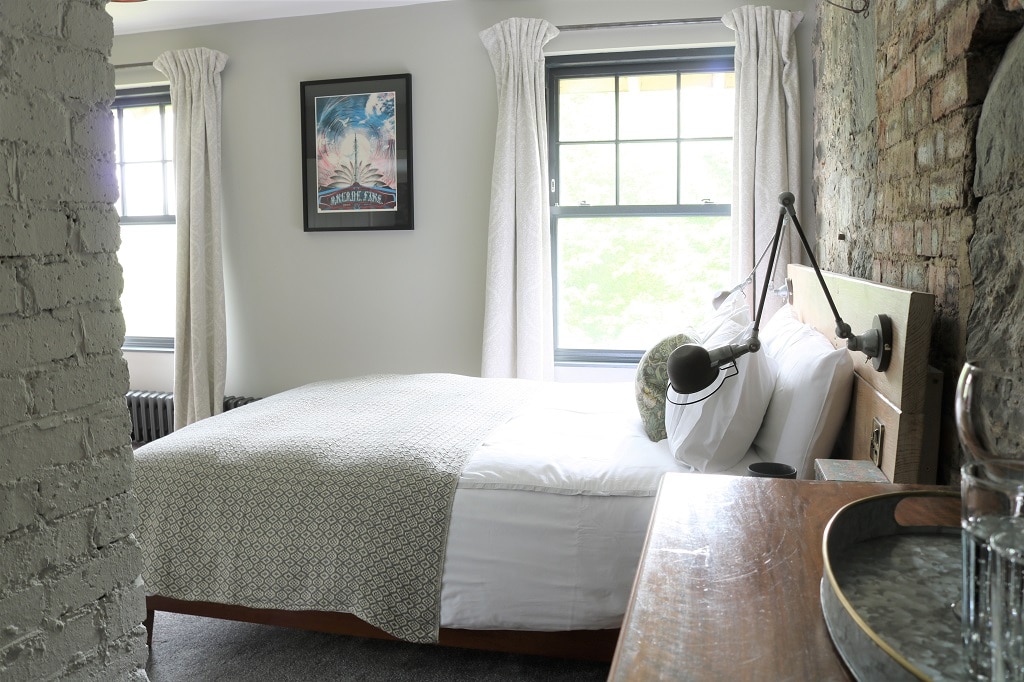 A bright room features a UK king size bed with a patterned throw and decorative pillows. Light enters through two windows, framed by soft curtains. A vintage style bedside lamp and wooden table complement the brick wall, enhancing the eclectic decor.
