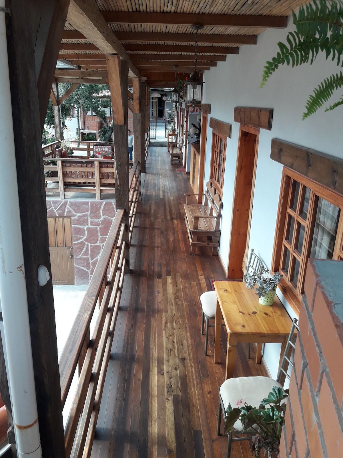 A wooden corridor is lined with rustic seating, featuring small tables and potted plants. Natural light filters through the open spaces, highlighting the warm tones of the wooden beams and flooring. Doorways to individual rooms can be seen along the passage.