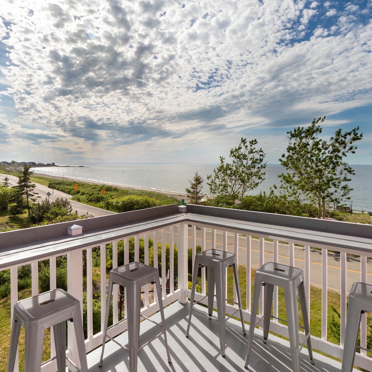 A spacious balcony features four metal bar stools, offering a panoramic view of the nearby beach and ocean. A cloud-filled sky reflects light over the water, and lush greenery lines the pathway along the coast, enhancing the serene atmosphere.