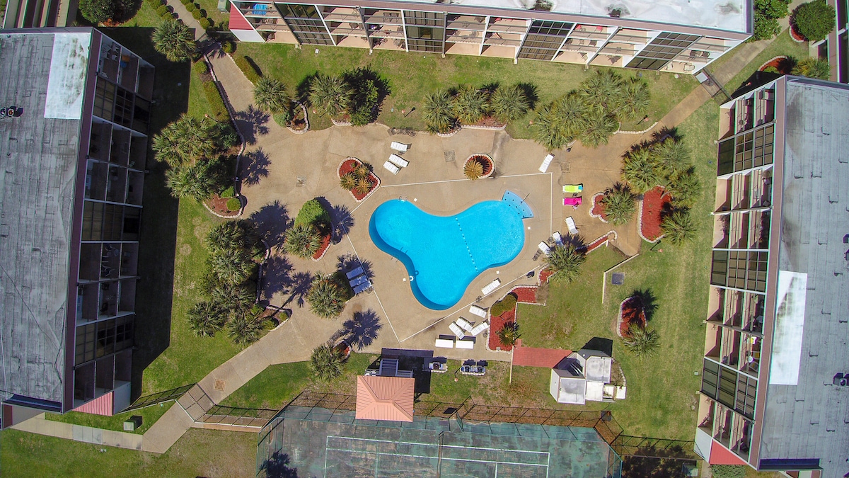 An aerial view of the property showcases a well-maintained in-ground pool surrounded by landscaped greenery and lounge chairs. Several buildings feature balconies overlooking the pool area, while a tennis court is visible in the bottom portion of the image.