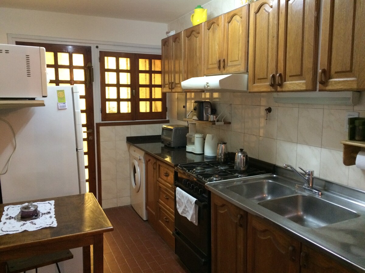 The kitchen area is equipped with wooden cabinets and a black countertop. A stainless-steel sink is positioned beside a gas stove with an oven. Appliances, including a microwave and toaster, are visible. A washing machine is located near a door leading to the outside. Light filters through the window with grid design.