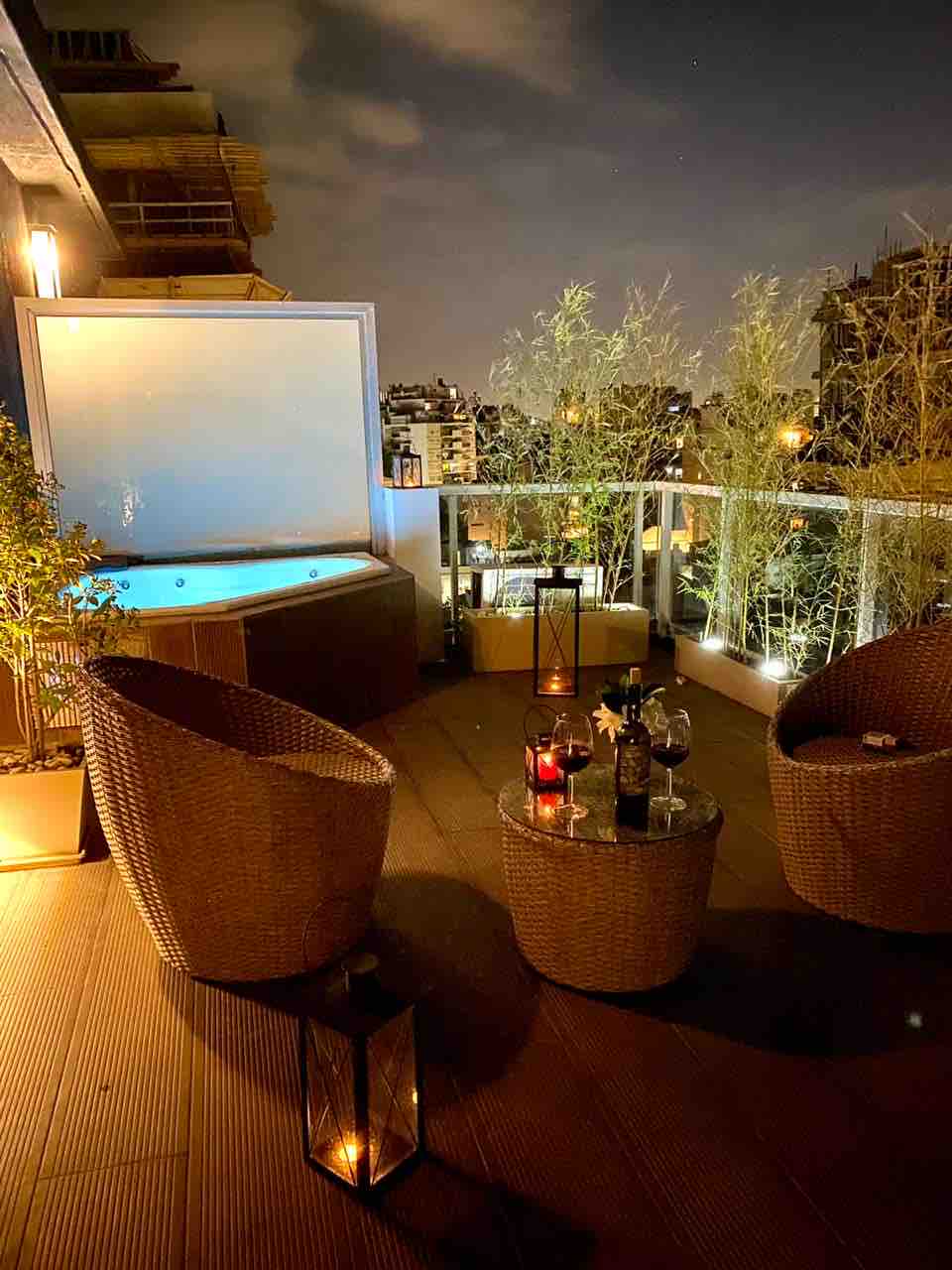 A private terrace is illuminated at night, featuring a Jacuzzi enclosed by a low wall. Two woven chairs and a round table are positioned under soft lighting, complemented by decorative plants. A tranquil city view enhances the serene setting.