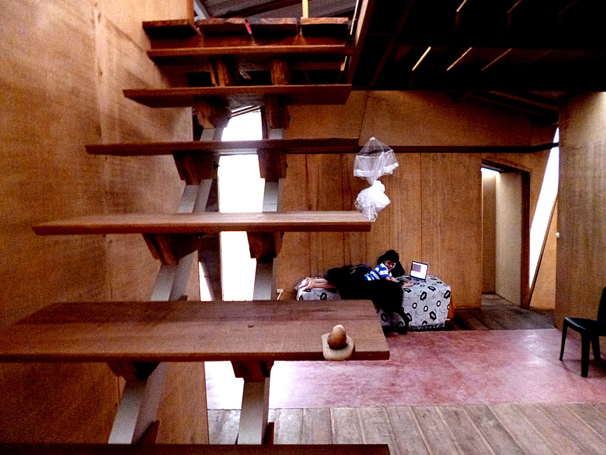 A wooden staircase with flat steps leads to an upper level. In the lower area, a person is seated on a patterned couch, using a laptop. Natural wood walls provide a warm backdrop, with soft light filtering into the space.