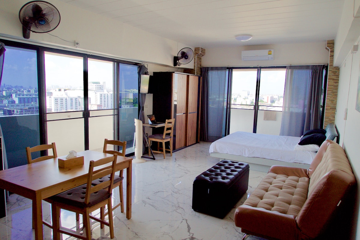 The open studio apartment features large windows allowing ample natural light and views of the surrounding area. A spacious bed is positioned alongside a sofa that can convert into an extra bed. A dining table and a flat-screen television are also included.