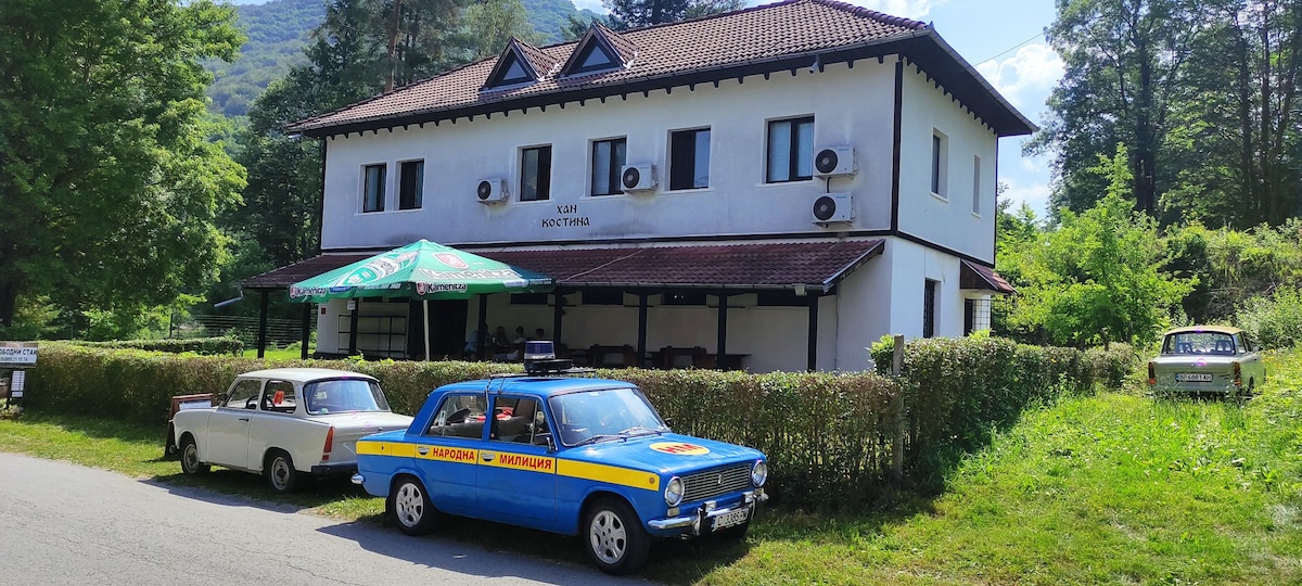 The two-story structure is surrounded by greenery, featuring multiple air conditioning units on its exterior. A blue vehicle with a yellow stripe is parked in front. An outdoor seating area is shaded by a canopy, inviting gatherings in a natural setting.