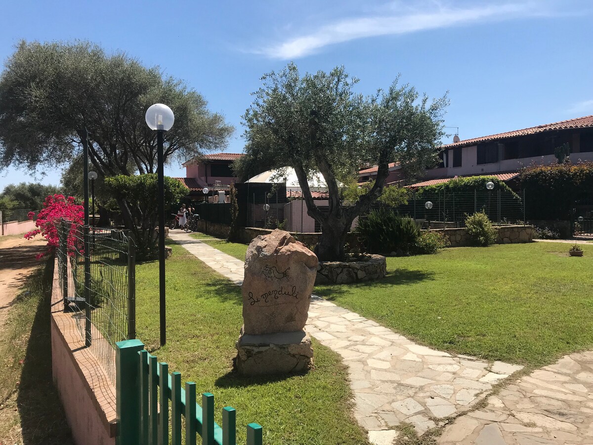 A landscaped garden area is showcased, featuring well-maintained greenery and stone pathways. Olive trees and flowering plants provide subtle color, while outdoor lighting fixtures line the path. In the background, a shaded seating area is visible under an awning.