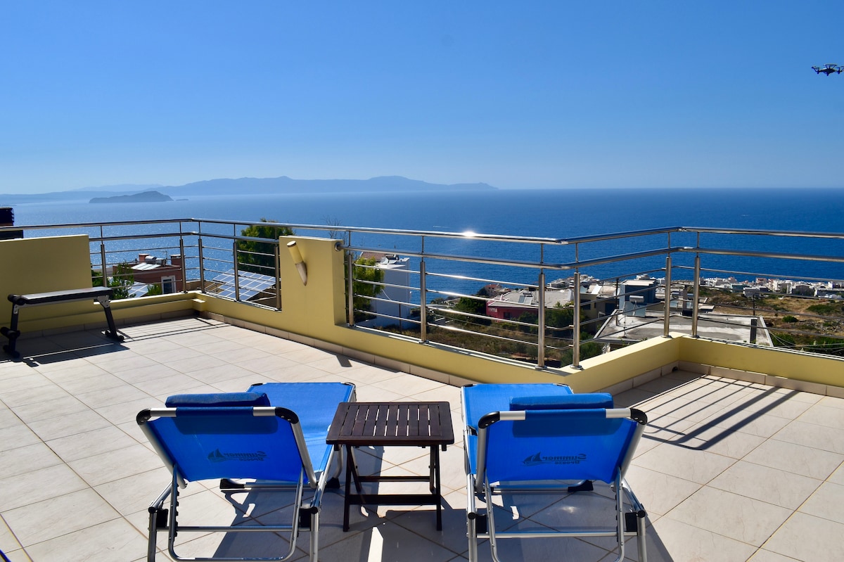 A spacious balcony offers a panoramic view of the sea and surrounding landscape. Two lounge chairs are placed alongside a small table, inviting relaxation. The clear sky enhances the serene atmosphere, while a railing ensures safety for children.