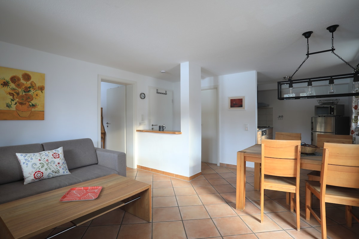 The living area features a gray sofa with an accent pillow, alongside a wooden coffee table. A dining table with four chairs is positioned beside a kitchenette, which includes stainless steel appliances. Light-colored walls and natural stone tiles create a warm atmosphere throughout the space.