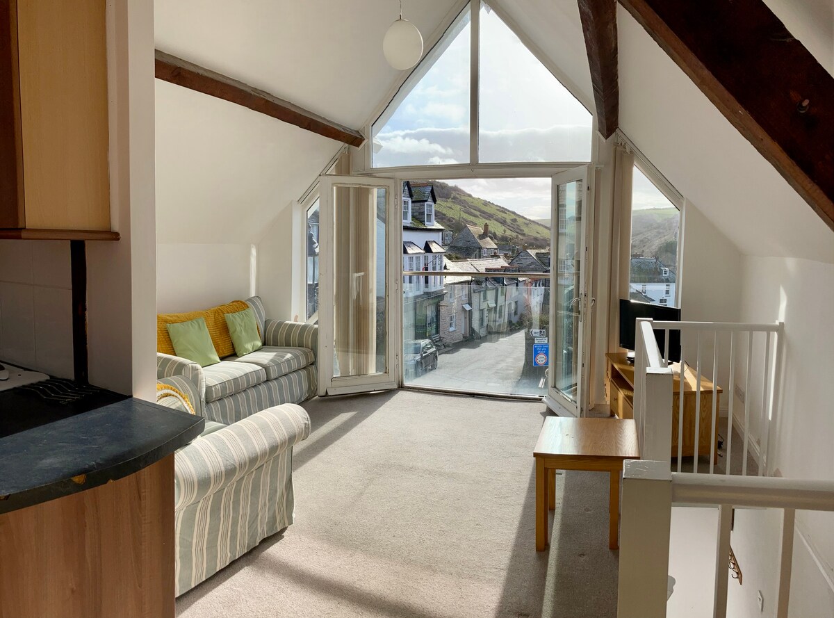 A large, airy lounge boasts high vaulted ceilings with exposed beams and double doors that open to a view of the village below. Two sofas are positioned in the space, accompanied by a small wooden coffee table, allowing for a comfortable area to relax.