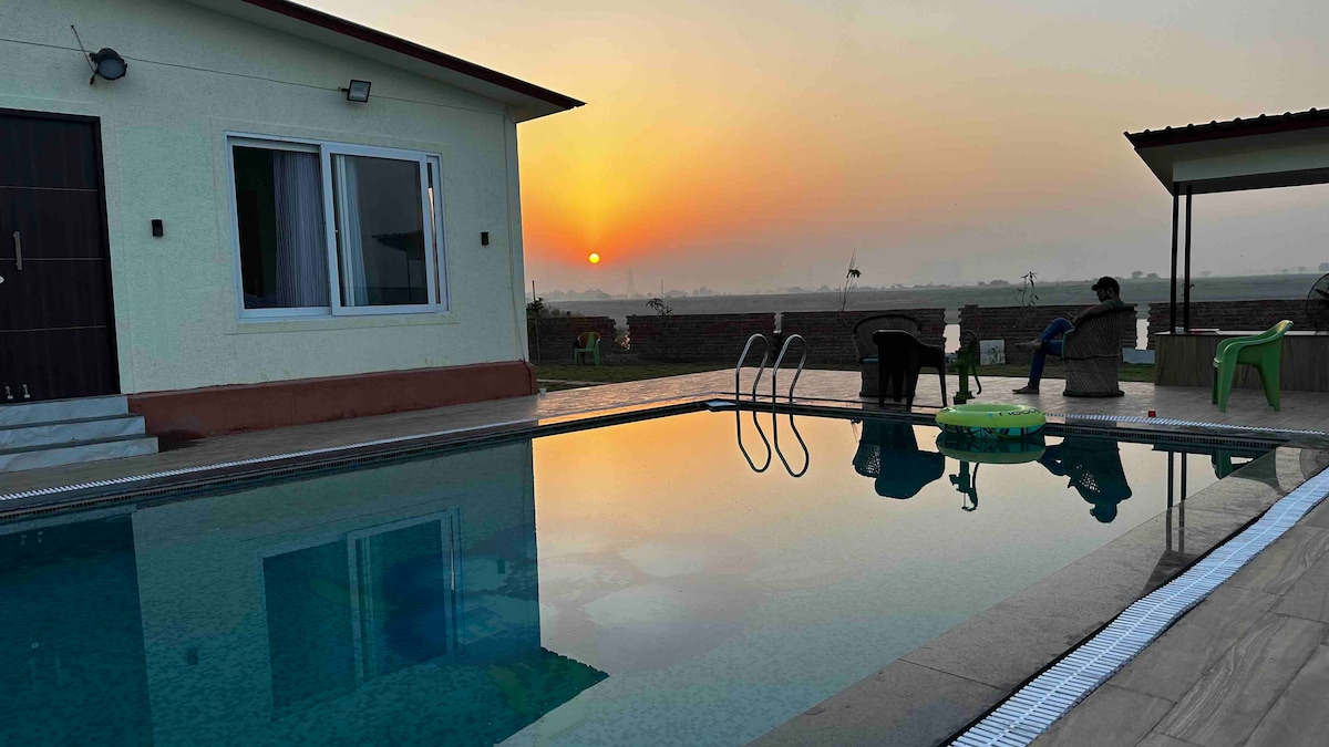 A tranquil swimming pool reflects the colors of a sunset, surrounded by a spacious patio. The modern building with large windows is positioned nearby, while green chairs are situated along the poolside. The serene landscape in the background enhances the peaceful ambiance.