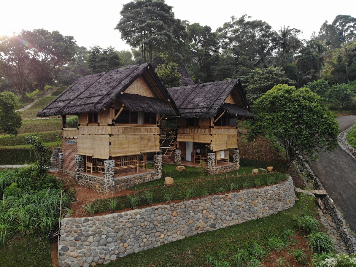 A bamboo structure comprising two units is showcased, set against a backdrop of lush greenery and gently rolling hills. The building features thatched roofs and stone accents, blending harmoniously with the natural surroundings. A winding path leads to the entrance, emphasizing the tranquil environment.