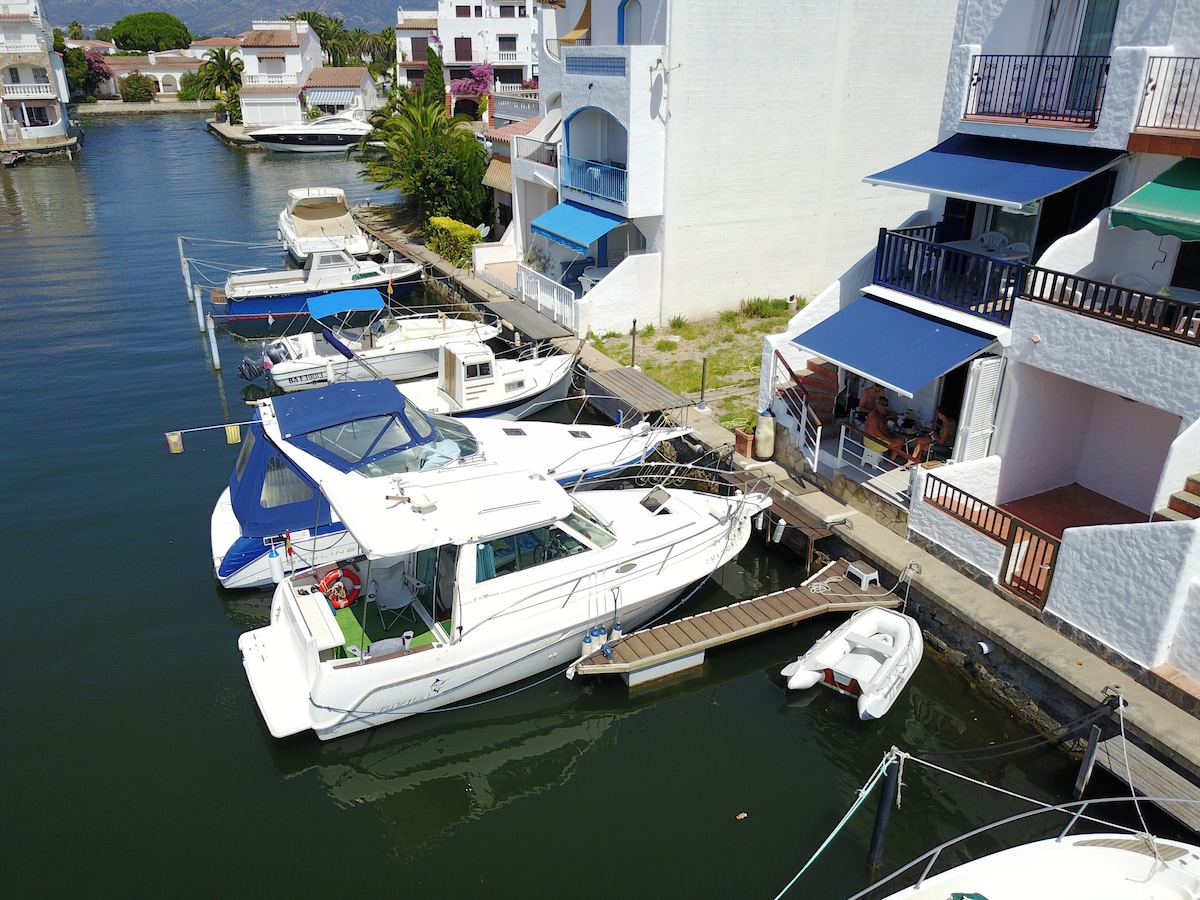 A view of a tranquil canal lined with boats, showcasing various vessels moored along the dock. Bright awnings can be seen above outdoor seating areas on the nearby building, offering shaded relaxation spots overlooking the water.