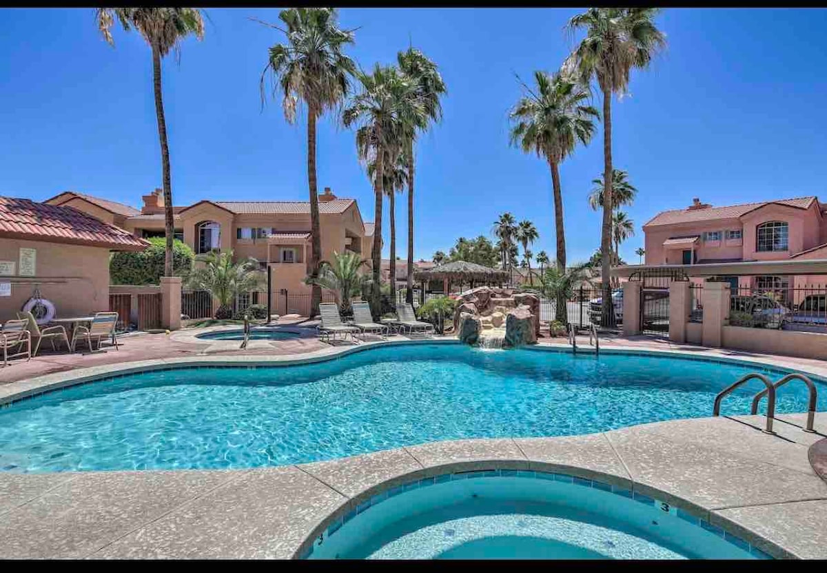 A spacious pool area is surrounded by palm trees, featuring a waterfall and a hot tub. Lounge chairs are arranged around the pool, inviting relaxation. The image displays a sunny day with clear blue skies, enhancing the tranquil ambiance of the condo community.