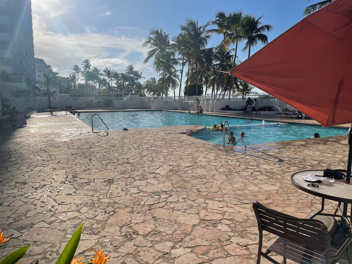 A spacious outdoor pool area is framed by palm trees under a bright sky. The clear blue water reflects sunlight, while a separate shallow section is visible. Lounge chairs and tables are arranged nearby, providing comfortable seating options for guests.