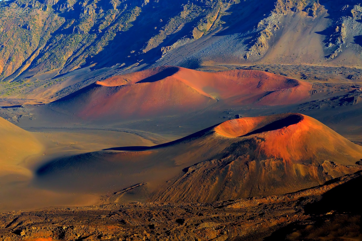 The image captures expansive volcanic craters with reddish hues contrasting against dark volcanic soil, framed by rugged mountainous terrain. The varying elevations and textures of the landscape create a dramatic visual, as sunlight casts shadows across the craters and surrounding areas.