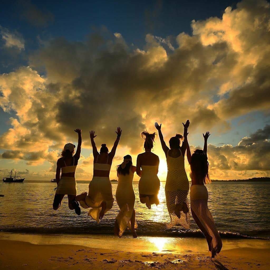 Silhouettes of six individuals are seen joyfully jumping against a backdrop of a golden sunset, with clouds softly illuminated by the setting sun. The tranquil water reflects the warm hues, creating a serene beach atmosphere.