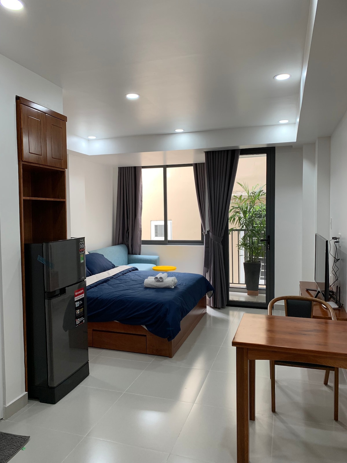 An open-plan studio apartment features a spacious bed with dark bedding and a light blue sofa. A small dining table is placed near the window, which offers a view of a balcony with greenery. A refrigerator and television are visible, contributing to the functional design.