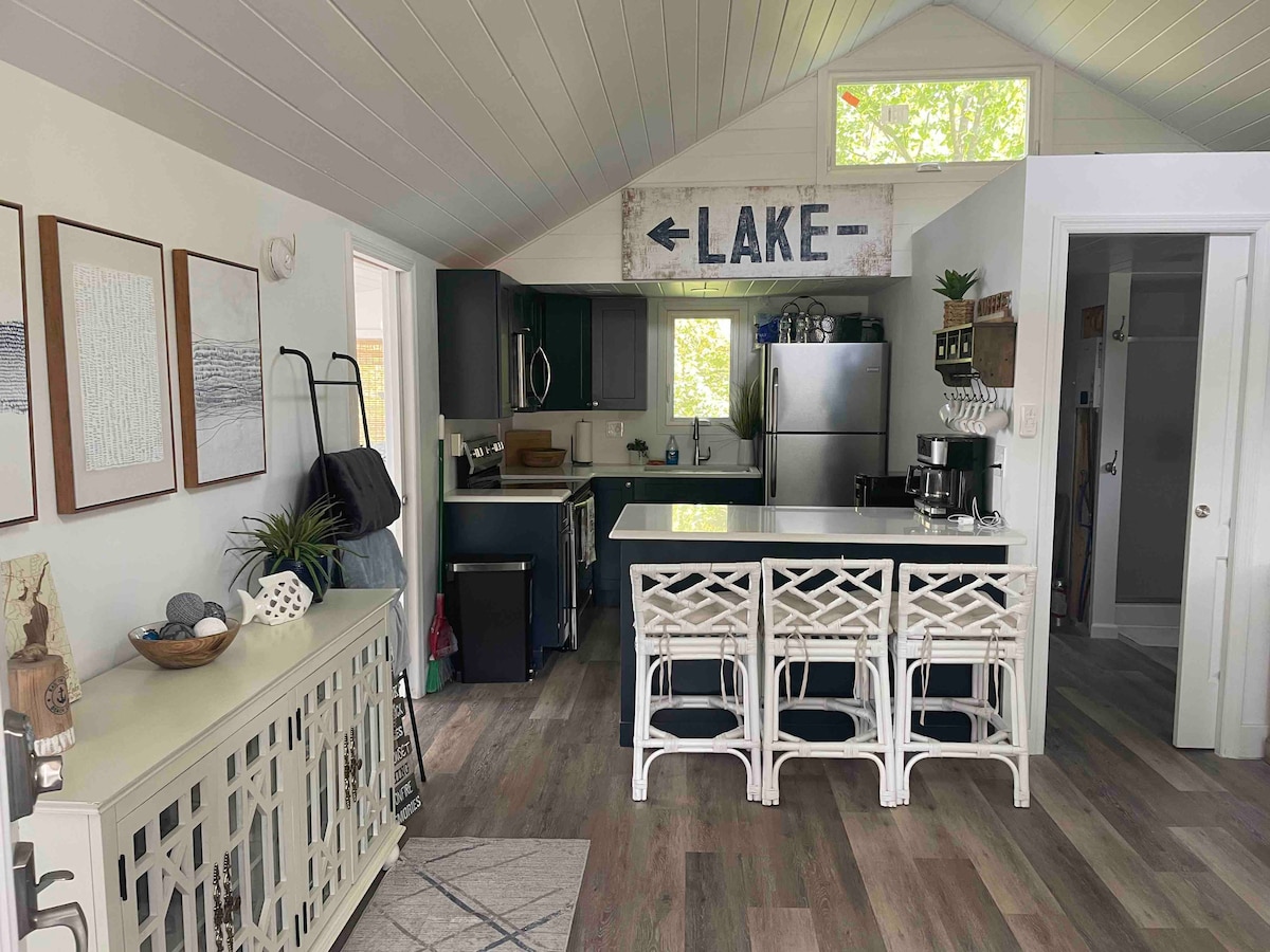 A modern kitchen is displayed with dark cabinetry and stainless steel appliances. A breakfast bar with white wicker stools provides seating. A sign labeled 'LAKE' is mounted on the wall, and natural light enters through the window, highlighting the open and airy design.