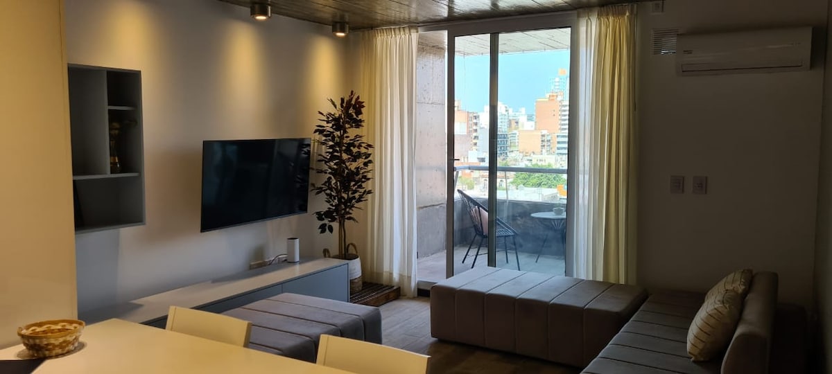 The living area showcases a modern design with a large smart TV mounted on the wall. Natural light streams through the floor-to-ceiling windows, leading to a balcony with outdoor seating. Soft, neutral tones are present in the furnishings, contributing to a comfortable atmosphere.