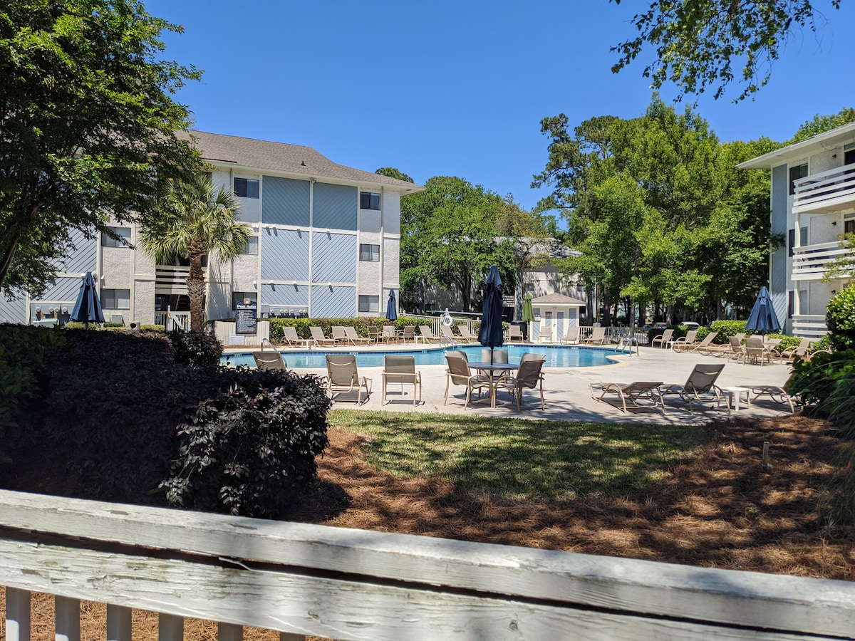 A spacious pool area is surrounded by inviting lounge chairs and umbrellas, set among well-maintained landscaping. Tall trees provide shade, enhancing the outdoor appeal. The villa buildings are visible in the background, reflecting a serene community atmosphere.