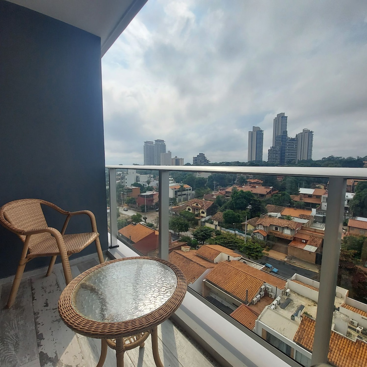 A balcony with two woven chairs and a round glass table offers a view of a green landscape and modern buildings. The urban skyline contrasts with the residential area below, while clouds drift across the sky, creating a calm atmosphere.