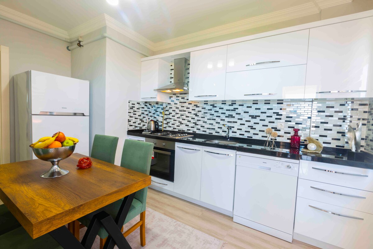 A modern kitchen is displayed, featuring white cabinetry and a glass-tiled backsplash. A wooden dining table accompanied by two green chairs is positioned in front. Stainless steel appliances, including a refrigerator and dishwasher, are included, complementing the open layout.