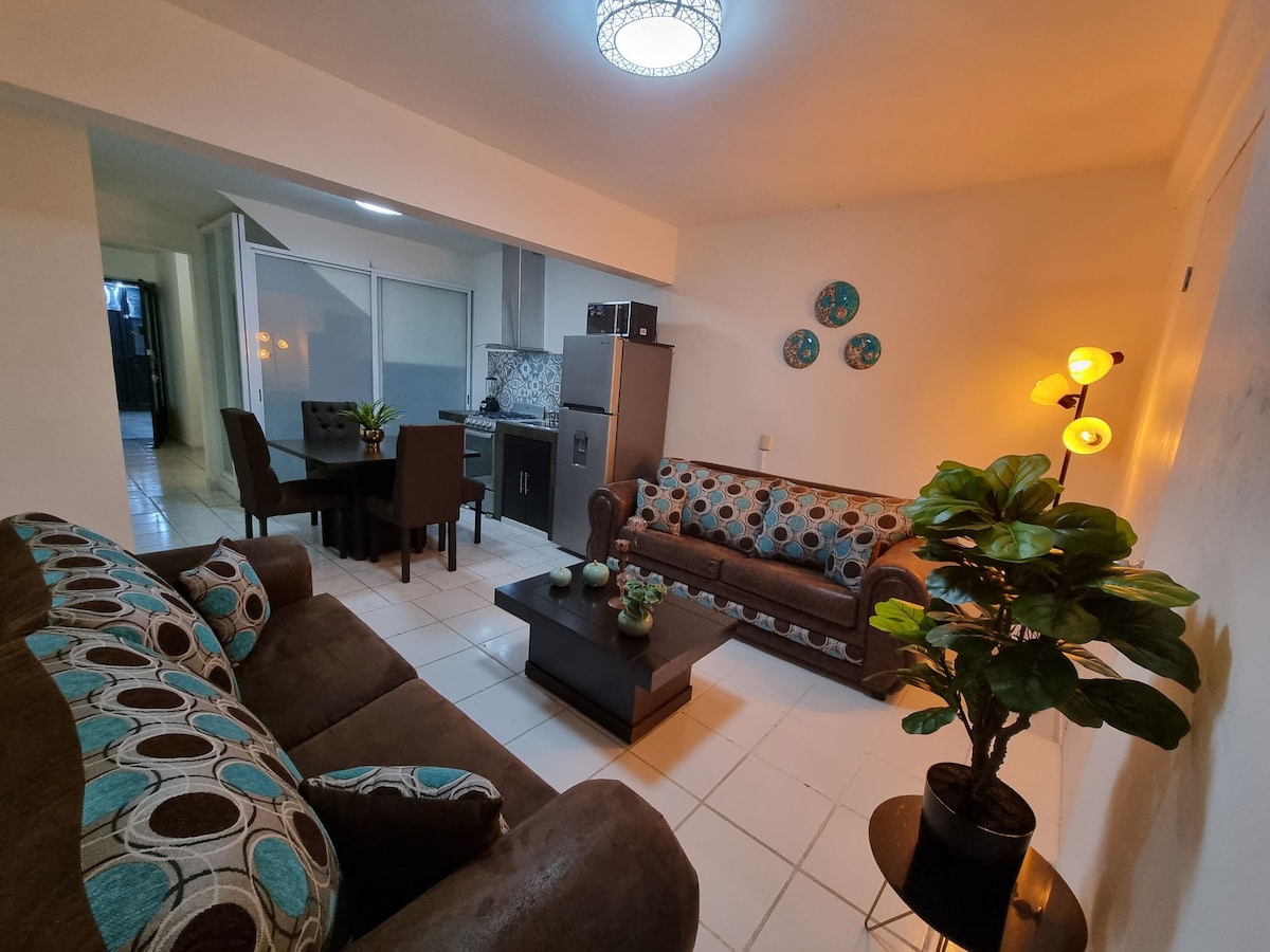 A cozy living area is highlighted by a comfortable brown sofa and armchairs featuring circular patterns. A dining table for four sits nearby, and a small plant adds a touch of greenery. Modern kitchen appliances and decorative wall elements are visible in the background.