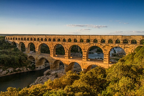 An ancient stone aqueduct spans above a river, characterized by multiple arches supported by sturdy columns. Lush greenery surrounds the base, while the sky showcases a fading light from dusk, casting warm tones onto the structure. The tranquil landscape is complemented by the flowing water beneath.