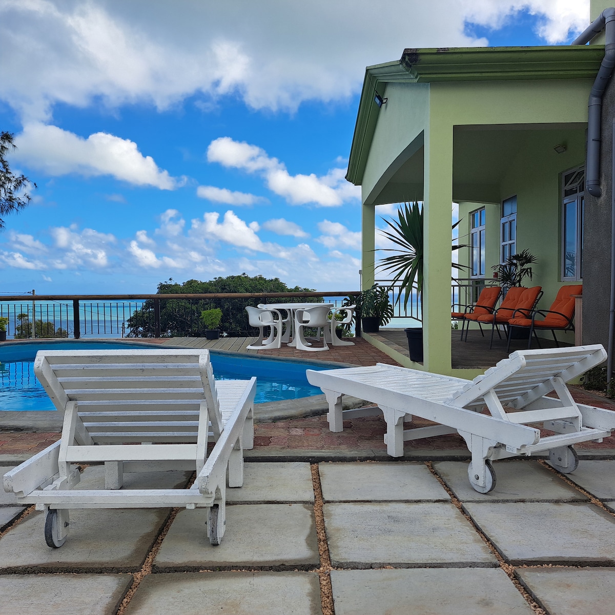 A private pool area is featured, with several white lounge chairs positioned on a stone deck. Nearby, a table and chairs are arranged on a patio overlooking the sea, framed by a vibrant sky dotted with clouds.