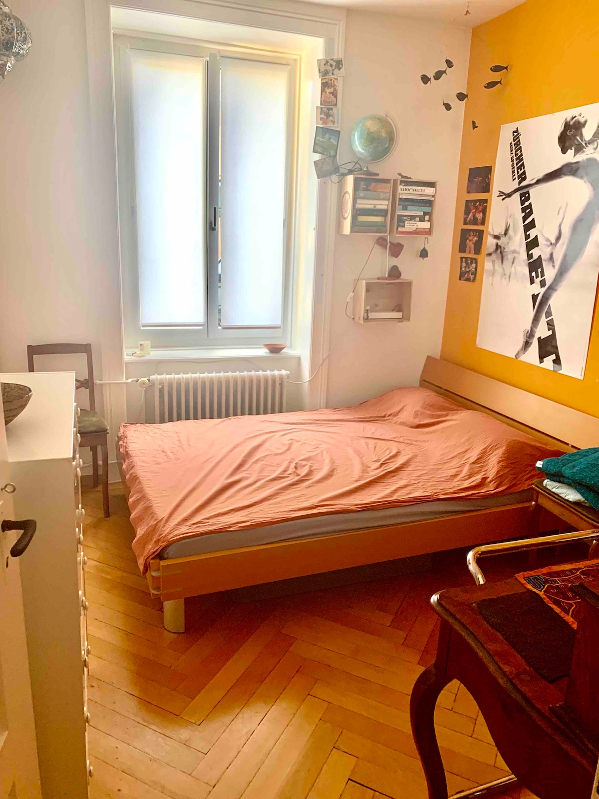 A compact bedroom features a wooden bed with a light-colored bedspread, positioned adjacent to a large window that allows natural light to enter. The honey-colored flooring complements the vibrant orange accent wall, which is adorned with wall art and photographs.