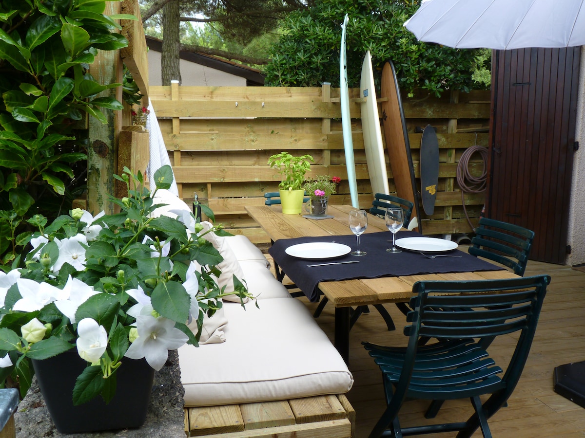An outdoor seating area is presented with a wooden dining table and green chairs, surrounded by lush greenery. A large white umbrella provides shade, while a soft cushion runs along one side. A potted plant and decorative flowers add a touch of color.