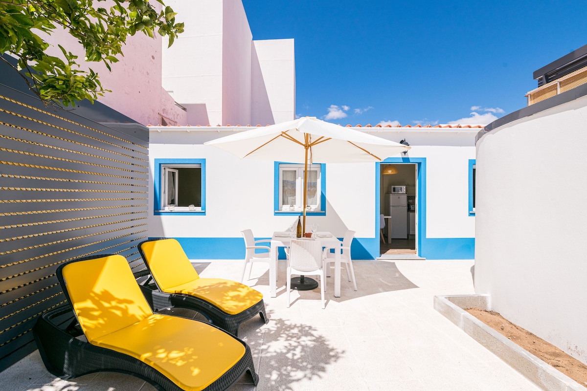 A sunlit outdoor terrace features two yellow lounge chairs and a white dining table with four chairs. An umbrella provides shade, and a light-colored wall contrasts with the vibrant blue trim of the structure, enhancing the inviting atmosphere.