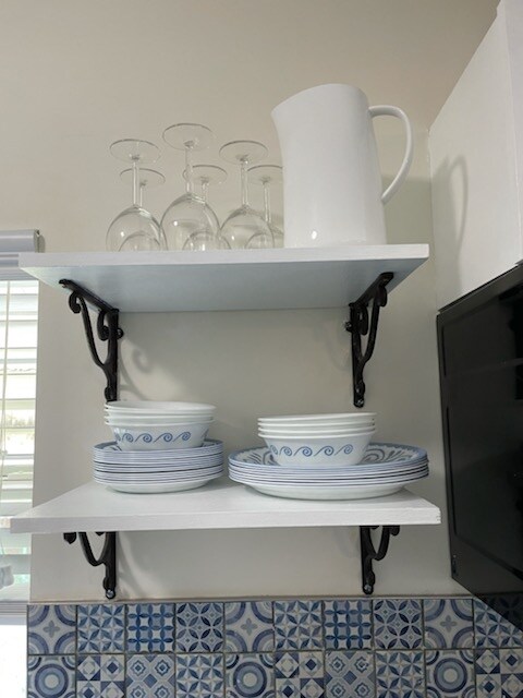 A set of white dishes with blue accents is displayed on white shelves, supported by decorative black brackets. Above, a clear glass pitcher stands next to several stemmed glasses. A colorful tiled backsplash with a geometric pattern complements the kitchen area.