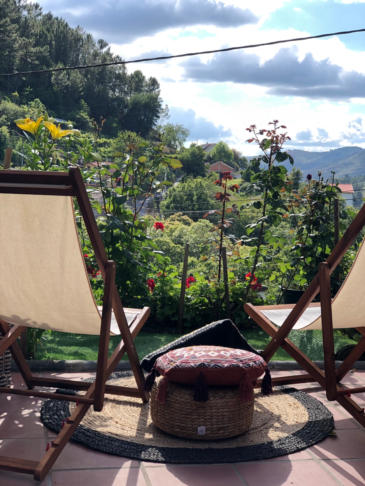 A serene outdoor seating area features two wooden chairs positioned to face a lush garden. Colorful flowers bloom among greenery, creating a vibrant backdrop. A round woven rug and a patterned pouf add texture to the space, while distant hills and clouds are visible under a partly sunny sky.