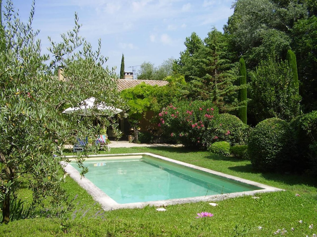 A serene swimming pool is set within a landscaped garden, surrounded by well-groomed greenery and colorful flowerbeds. A shaded area features a patio umbrella, adding to the tranquil atmosphere. Tall trees and hedges frame the area, contributing to the sense of privacy.