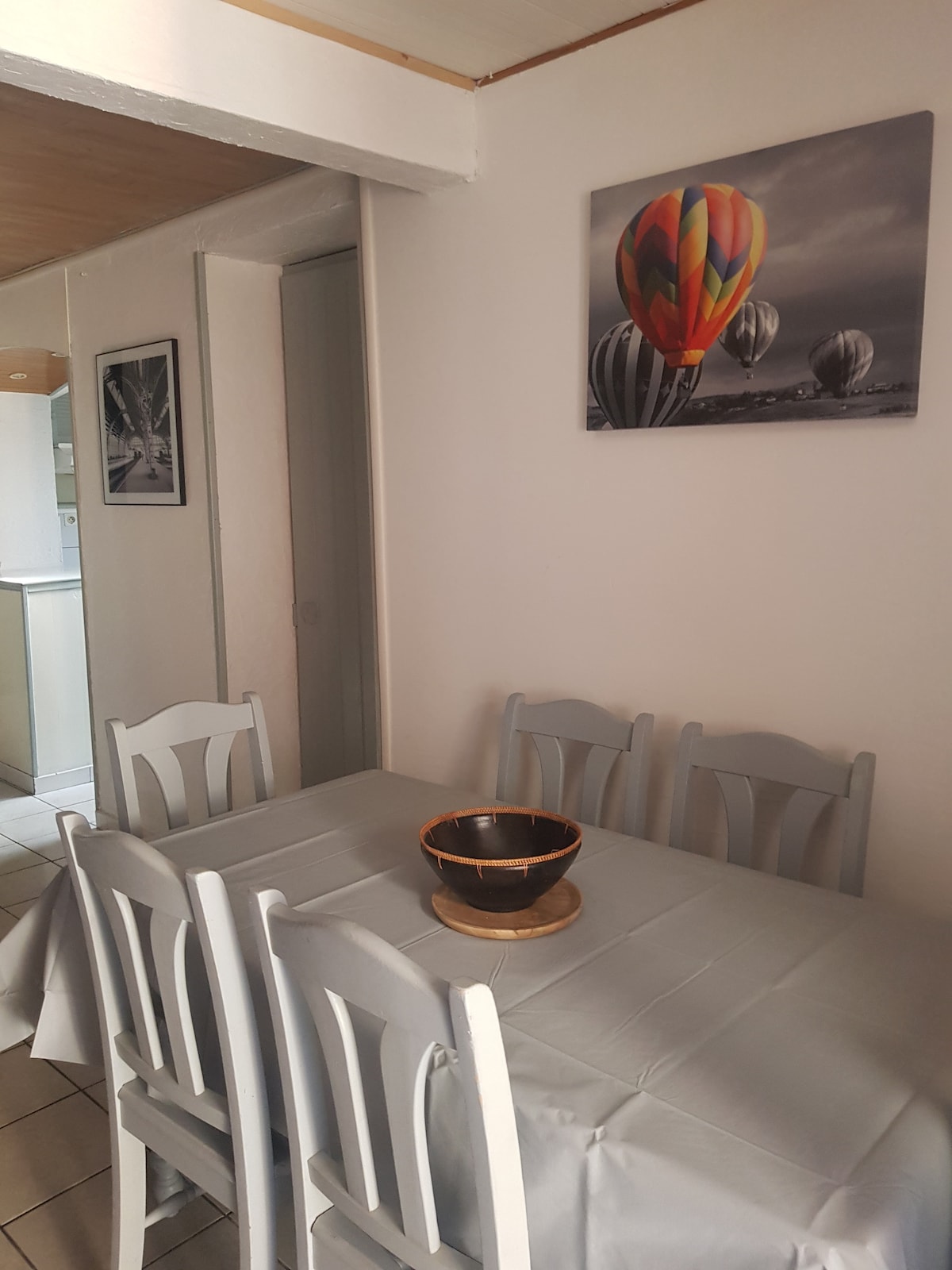 A dining area features a rectangular table set with a light grey tablecloth. Six light-colored chairs surround the table, and a wooden bowl sits in the center. A vibrant wall art piece depicting hot air balloons is mounted above the table, contributing to a cheerful ambience.