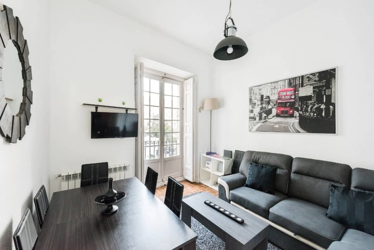 A bright living room features a gray sofa and a dark wooden dining table surrounded by six chairs. A wall-mounted television is positioned above a small shelf. Natural light floods the space through double doors that open to a balcony, enhancing the airy atmosphere.