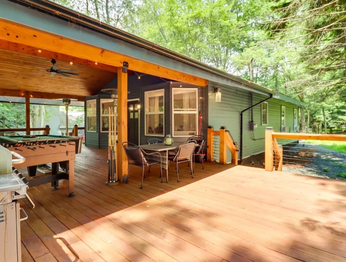 A spacious outdoor deck features a wooden floor and a ceiling fan, offering areas for seating with a dining table and chairs. The surroundings include trees providing a natural backdrop, enhancing the relaxed atmosphere of the cottage retreat.