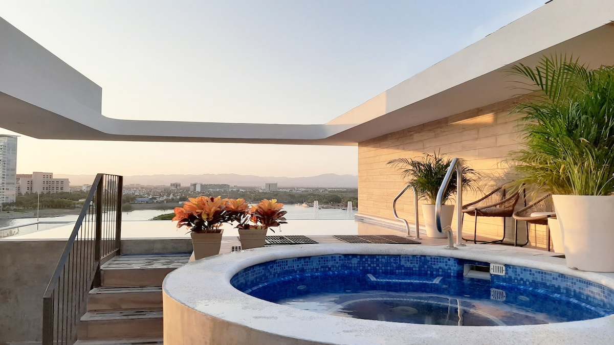 The rooftop area features a jacuzzi surrounded by potted plants and two comfortable chairs. A staircase leads to the lower level. The sunset view is visible in the background, creating a serene setting for relaxation.