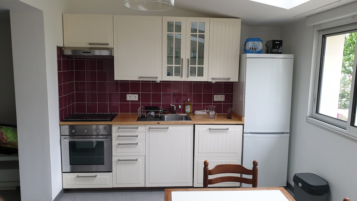 The kitchen features modern cabinetry with a wooden countertop and is complemented by burgundy tiled walls. Stainless steel appliances, including a refrigerator and oven, are present. A small dining table with a wooden chair is positioned nearby, and a window allows natural light to brighten the space.