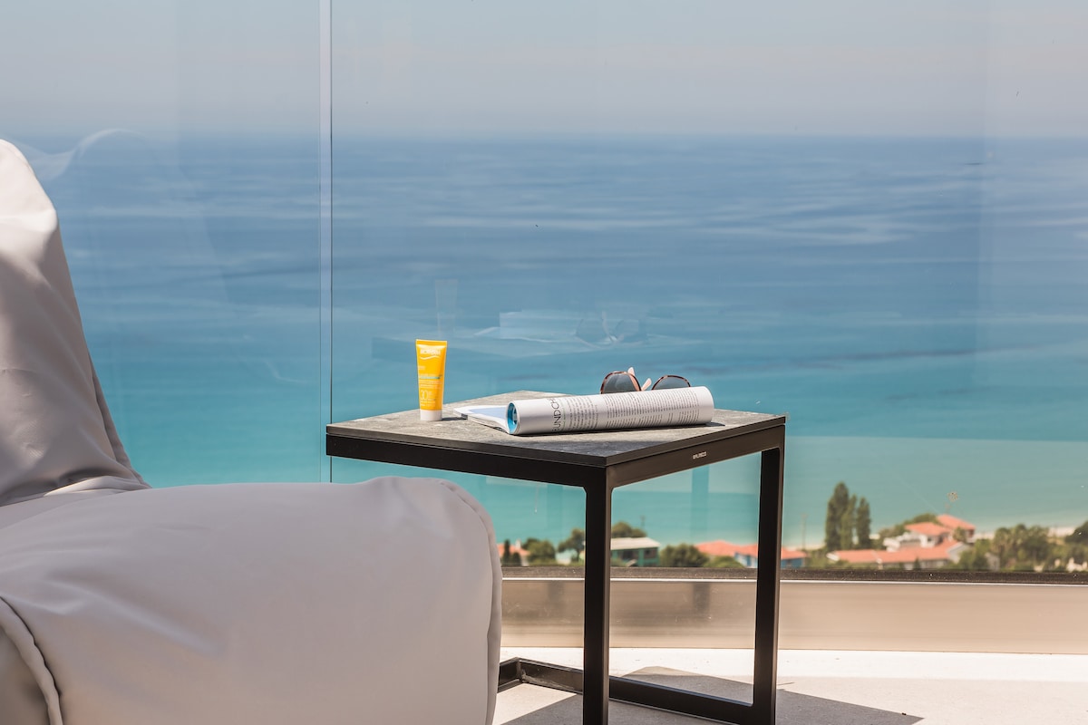 A modern outdoor seating area features a sleek table with a patterned magazine and a sunscreen bottle. The glass railing provides an unobstructed view of the calm blue sea and the distant coastline, enhancing the relaxing environment.