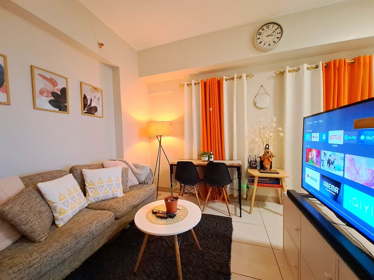 A cozy living area features a comfortable sofa adorned with patterned pillows. A small round table is positioned nearby, accompanied by two chairs. Natural light filters through orange and white curtains, while a wall clock adds a functional touch. A television displays various streaming options.