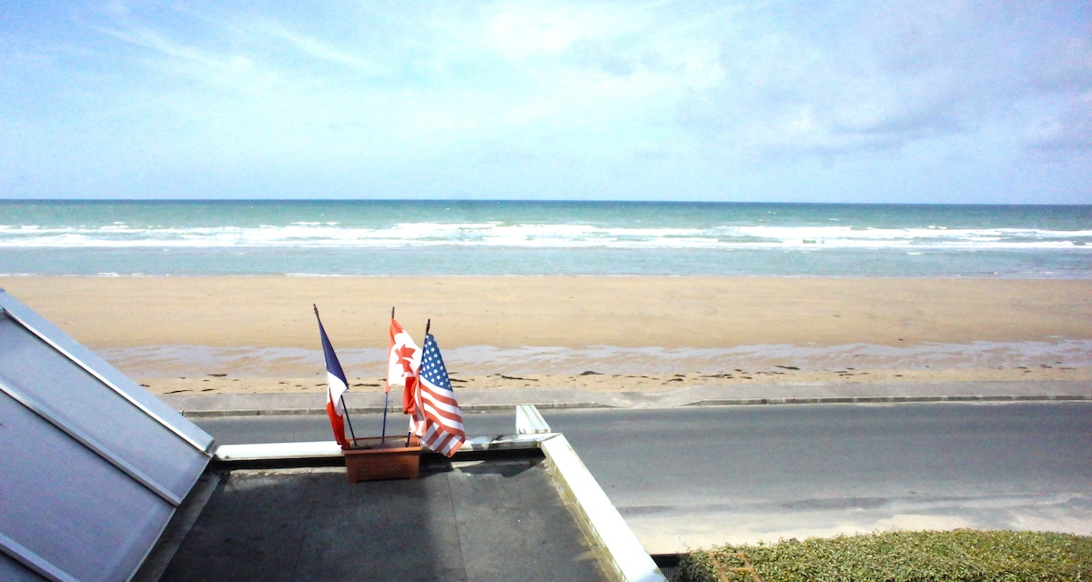 The view features a wide sandy beach stretching towards the horizon, with gentle waves lapping at the shore. Several flags, including the French, Canadian, and American flags, are displayed prominently in the foreground, suggesting a welcoming coastal ambiance.